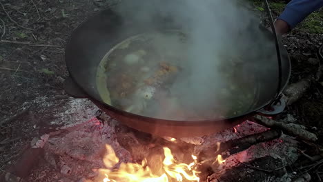Cooking-steaming-stew-on-top-of-blazing-flame-in-woodland,-slow-motion-view