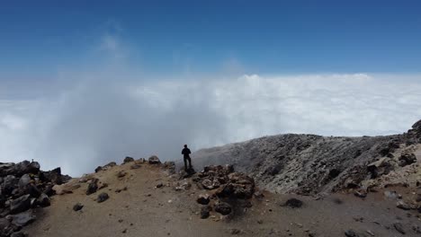 Man-stands-on-edge-of-volcano-crater-looking-at-sky-of-clouds-below