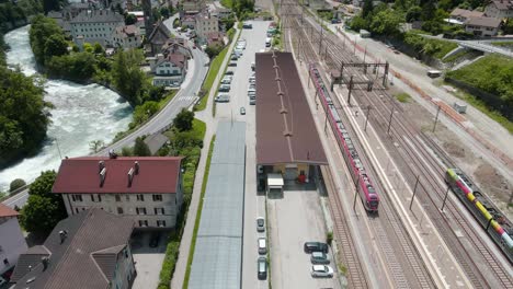Establishing-shot-of-train-station-near-river-stream,-small-town-and-green-vegetation---Cinematic-Drone-View