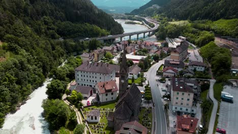 Vista-Aérea-De-Un-Pequeño-Pueblo-Italiano-Entre-Montañas-Verdes,-Cerca-Del-Arroyo-Azul-Del-Río-Eisack,-Un-Puente-Alto-Y-Una-Estación-De-Tren---Hermosa-Vista-Panorámica