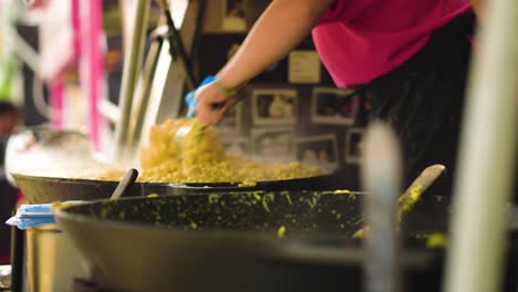 outdoor-cooking-of-mac-and-cheese-in-a-massive-frying-pan-at-the-festival-the-chef-is-moving-and-stirring-the-cheesy-pasta-getting-in-ready-for-customers-to-stay-fresh-wearing-one-glove