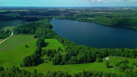 lake-flyover-aerial,-tilt-up-to-cloudy-sky,-idyllic-rural-area,-green-meadows,-big-water-pond,-blue-sky-with-peaceful-clouds,-drone-landscape-overview-of-the-countryside