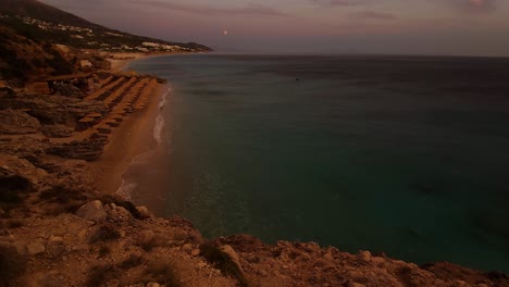 Seaside-of-Ionian-sea-in-Albania-with-beautiful-beaches-and-resorts-on-hills-at-golden-hour,-full-moon-on-purple-sky