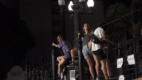 Un-Grupo-De-Mujeres-Jóvenes-Por-La-Noche-Trepa-Por-Las-Puertas-De-Un-Edificio-Público-Para-Tomar-Fotografías-Grupales,-Una-Bebe-Alcohol