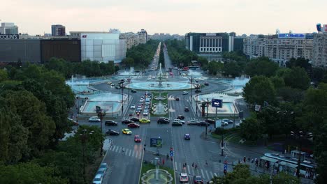 Vista-Aérea-Sobre-La-Plaza-Unirii-Con-Las-Famosas-Fuentes-De-Agua-En-El-Marco-Principal,-Al-Anochecer,-Bucarest,-Rumania,-Tráfico-Pesado,-Autos-Pasando,-Fuentes-De-Agua-Azul