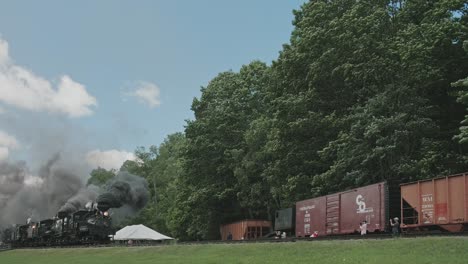 Ein-Blick-Von-Unten-Auf-Eine-Dampflokomotive-Mit-Fünf-Verbundenen-Dampflokomotiven,-Die-Sich-Nähert,-Viel-Rauch-Und-Dampf-Ausstößt-Und-An-Einem-Sonnigen-Sommertag-An-Güterwaggons-Vorbeifährt
