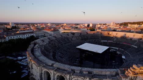 Toma-Aérea-Con-Drones-Del-Famoso-Estadio-De-Nimes,-Una-Ciudad-En-El-Sur-De-Francia,-Vista-Frontal-E-Interior-Con-La-Puesta-De-Sol-Fuera-De-Cámara-Iluminando-La-Ciudad,-Calles-Tranquilas-Y-Algunos-árboles-Y-Pájaros.