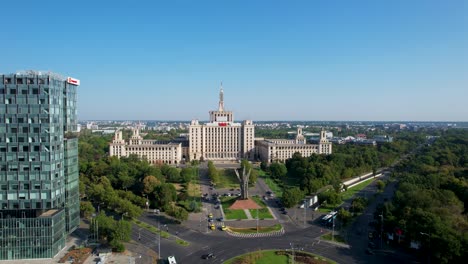 Aerial-View-Of-The-House-Of-The-Free-Press-Building-In-Bucharest,-Romania,-Passing-By-Tall-Office-Buildings,-Surrounded-By-Green-Vegetation