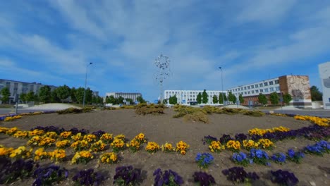 Windspinner-Kinetische-Kunstskulptur-Aus-Rostfreiem-Metall-In-Einem-Mit-Blumen-Bepflanzten-Kreisverkehr