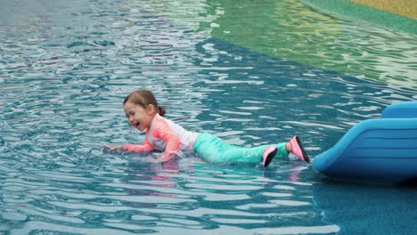 Happy-Little-Toddler-in-Swimsuit-Refreshing-Lying-in-Shallow-Water,-Gets-Up,-Playful-Girl-Jumps-and-Smiles-at-Water-Park-Playground---slow-motion