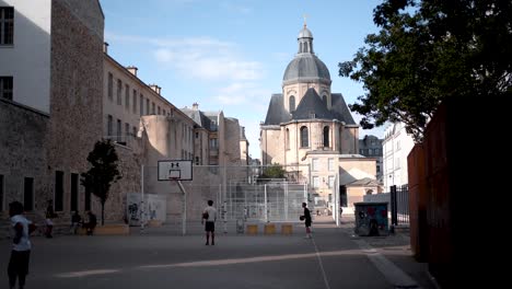 Niños-Jugando-Baloncesto-En-La-Cancha-Del-Patio-Trasero-De-París:-Vibraciones-De-Vecindario-Con-La-Iglesia-Local-A-La-Vista,-Deportes-Y-Comunidad-En-Un-Entorno-Urbano