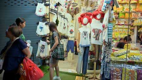 Choosing-some-possible-souvenirs-of-shirts,-pants,-and-some-delicacies-from-a-local-vendor-in-Chatuchak-Weekend-Night-Market,-in-Bangkok,-Thailand