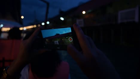 Un-Turista-Tomando-Vídeos-Y-Fotografías-Mientras-Pasa-Por-Algunas-Casas-Locales,-Tiendas-De-Souvenirs-Y-Restaurantes-En-El-Mercado-Flotante-De-Amphawa-En-Samut-Songkhram,-Tailandia.