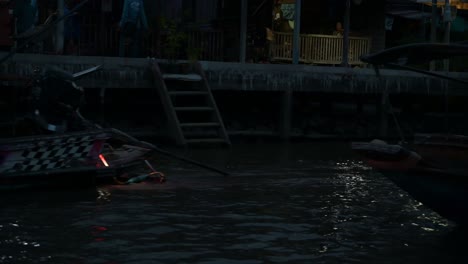 Moving-from-the-left-to-the-right-side-of-the-frame,-a-wooden-boat-filled-with-local-and-foreign-tourists-are-on-a-night-tour-at-Amphawa-Floating-Market-in-Samut-Songkhram,-Thailand