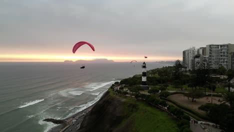 Aerial-travelling-shot-showing-a-pair-of-paragliders-flying-in-the-coast-near-a-lighthouse-at-sunset-time-in-the-middle-of-a-park-next-to-some-buildings-with-the-ocean-and-an-island-in-the-background