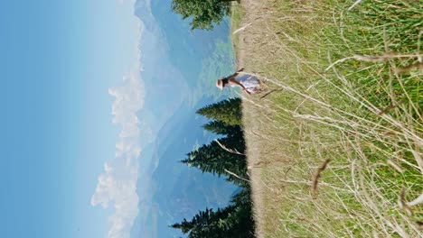 Vertical-shot-of-happy-woman-with-white-skirt-jumping-in-green-grass-field-during-sunny-day---Beautiful-mountain-range-and-valley-in-background---Slow-motion
