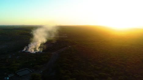 Smoke-plume-rises-from-burn-pit-at-sunset,-golden-hour-glow-spreads-across-shrub-landscape,-aerial-trucking-pan