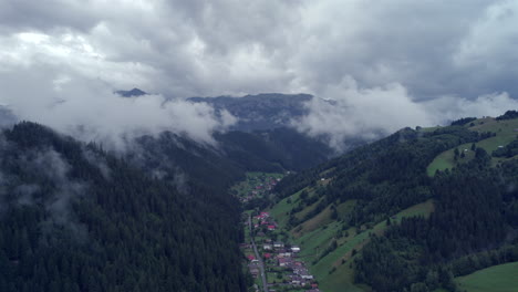 Aerial-view-along-a-valley-with-a-mountain-resort-village,-high-mountains-with-fir-forests-and-very-low-clouds