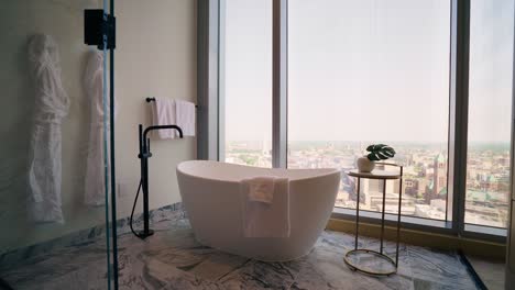 Wide-shot-of-modern-white-free-standing-bath-tub-in-high-rise-bathroom-with-large-windows-and-a-city-view