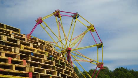 revealing-big-wheel-roller-coaster-yellow-and-red-colour-behind-stack-of-pallets-festival-park-scenery-partly-cloudy-summer-vibe-holiday-camping-trees-in-the-background