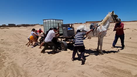 Eine-Gruppe-Westlicher-Kinder-Und-Erwachsene-Hilft-Arabischen-Menschen-Dabei,-Pferdekarren-Mit-Wasser-Aus-Dem-Sand-Zu-Entfernen