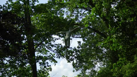 Skulptur-Eines-Nackten-Mannes,-ähnlich-Wie-Peter-Pan,-Der-Sich-Am-Himmel-Zwischen-Bäumen-Dreht,-Versteckt,-Ausgebreitete-Arme,-Gebeugte-Knie,-Unheimlich-Aussehend,-Mystischer-Thriller-Horror,-Teilweise-Bewölkt,-Näher-Heranzoomend,-Filmisch