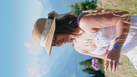 Portrait-shot-of-young-brunette-girl-with-hat-smelling-flowers-on-slope-of-alps-in-summer,-close-up