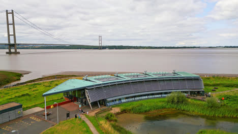 Aerial-footage-showcases-the-Waters'-Edge-Country-Park-and-Visitor-Centre,-gracefully-positioned-beside-the-Humber-Bridge