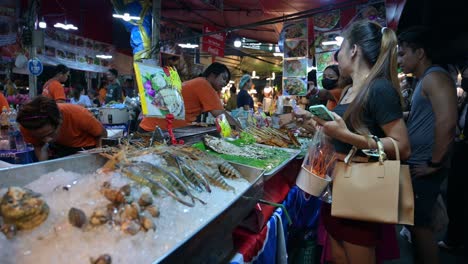 En-El-Lado-Derecho,-Se-Puede-Ver-A-Los-Clientes-Esperando-Sus-Pedidos-De-Mariscos-Y-Carne-De-Cerdo-Y-Pollo-A-La-Parrilla-De-Los-Vendedores-Locales-Dentro-Del-Mercado-Nocturno-De-Fin-De-Semana-De-Chatuchak-En-Bangkok,-Tailandia.