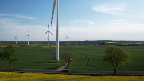 Rückwärtsantenne-In-Der-Nähe-Riesiger-Windkraftanlagen-Auf-Landwirtschaftlichen-Feldern-Und-In-Der-Nähe-Eines-Gelben,-Reifenden-Rapsfeldes