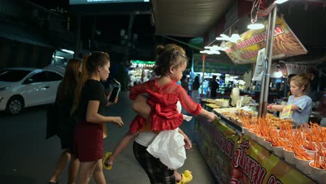 Tres-Mujeres-Y-Un-Bebé-Buscan-Comprar-Algunas-Frutas-Y-Bocadillos-Para-Amigos-Exhibidos-En-Un-Puesto-De-Comida-En-El-Mercado-Nocturno-De-Fin-De-Semana-De-Chatuchak-En-Bangkok,-Tailandia