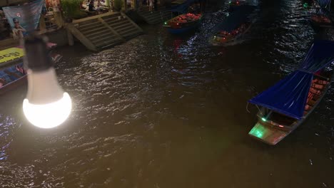 Moviéndose-De-Arriba-A-La-Derecha-A-Abajo-A-La-Izquierda,-Se-Puede-Ver-Un-Barco-Navegando-Por-El-Canal-Del-Mercado-Flotante-De-Amphawa,-Samut-Songkhram,-Tailandia,-Con-Una-Bombilla-Iluminando-El-Camino.