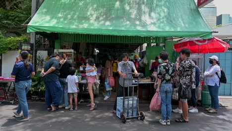 A-famous-roadside-restaurant,-serving-mostly-Northeastern-Thai-food,-packed-with-customer-up-front-while-some-motorcycles-are-passing-by-in-this-small-street-in-the-city-of-Bangkok,-Thailand