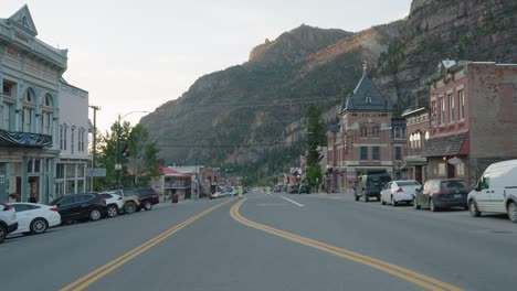 Looking-Down-Main-Street-In-Ouray,-Colorado-With-Traffic-Going-Past-During-Golden-Hour-With-San-Juan-Mountains-In-The-Background