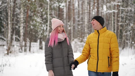 A-man-in-a-yellow-jacket-and-a-girl-in-a-hat-and-scarf-walk-through-the-winter-forest-during-a-snowfall-laughing-and-smiling-at-each-other-at-Christmas-in-slow-motion