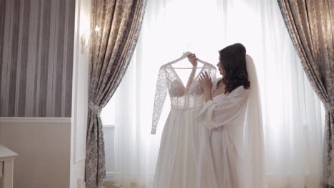 Bride-in-lingerie-standing-with-her-wedding-dress-at-home-near-window,-white-boudoir-dress-and-veil