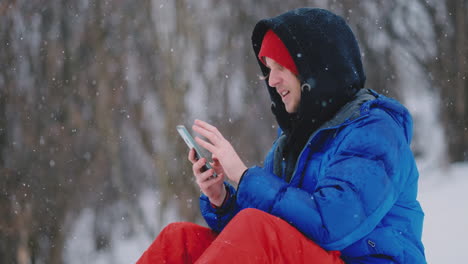 Ein-Männlicher-Snowboarder,-Der-Im-Schnee-Sitzt,-Fotografiert-Mit-Dem-Handy-Eine-Wunderschöne-Resortlandschaft-Für-Soziale-Netzwerke.-Resort-Blogger