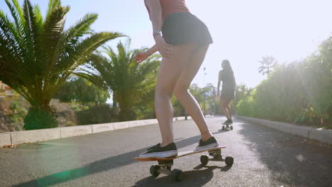 Zwei-Mädchen-Im-Skatepark-Fahren-Im-Sonnenlicht-Auf-Longboards-Die-Strecke-Entlang,-Freuen-Sich-Und-Lachen-In-Zeitlupe.-Steadicam-Rückansicht