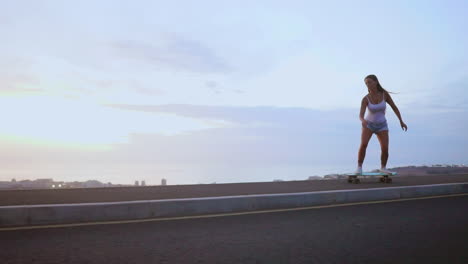With-the-sun-descending,-a-stunning-and-trendy-skateboarder-elegantly-skates-in-shorts-along-a-mountain-road-at-sunset,-with-the-mountains'-stunning-panorama-in-slow-motion