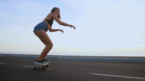 Enjoying-the-serene-sunset,-a-woman-skateboarder-rides-along-a-road-in-slow-motion,-with-mountains-and-a-stunning-sky-as-the-backdrop.-She-wears-shorts