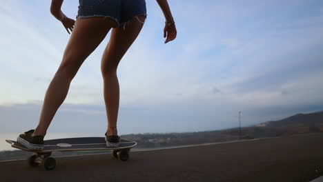 Captured-in-slow-motion,-a-stunning-young-skateboarder,-dressed-stylishly-in-shorts,-rides-her-board-along-a-mountain-road-at-sunset,-revealing-a-stunning-mountain-view