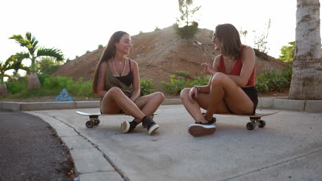 In-the-gentle-sunset-light,-two-girls-find-a-cozy-spot-at-the-skate-park,-sitting-on-their-boards,-immersed-in-conversation,-and-sharing-joyful-laughter.-A-longboard-chat-filled-with-smiles