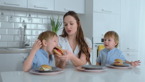 A-young-beautiful-mother-in-a-white-dress-with-two-children-is-smiling-and-eating-fresh-burgers-in-her-kitchen.-Happy-family-homemade-food-healthy-foods