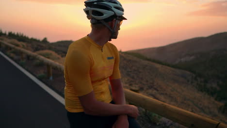 On-mountain-peak,-athlete-in-yellow-t-shirt,-helmet,-and-gear-relaxes-on-bike,-taking-in-mountain-view-and-sunset-after-training