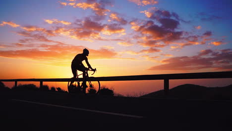 Silhouette-The-athlete-on-a-bike-is-showcased-in-slow-motion-as-he-rides-a-mountain-serpentine,-relishing-the-island's-view,-symbolizing-a-dedication-to-a-healthy-lifestyle-during-sunset