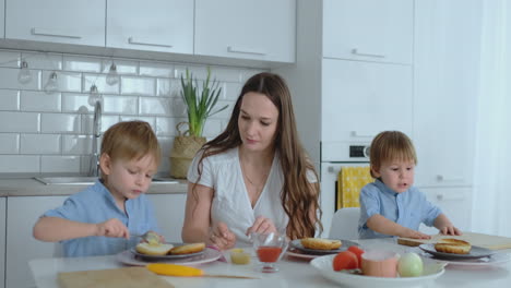 Mutter-Mit-Zwei-Kindern-In-Der-Küche-Am-Tisch-Und-Bereitet-Burger-Zum-Mittagessen-Zu