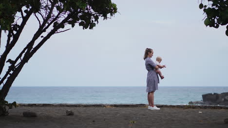 Amidst-the-beauty-of-the-sea-and-beach,-a-touching-portrait-showcases-a-mother-kissing-her-daughter's-cheek.-A-vacationing-family-shares-moments-of-joy,-and-the-mom-lovingly-hugs-her-toddler