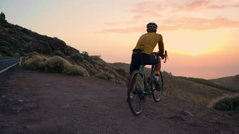 Post-training,-athlete-in-yellow-t-shirt,-helmet,-and-gear-sits-on-bike-on-mountain-peak,-enjoying-view-of-mountains-and-sunset