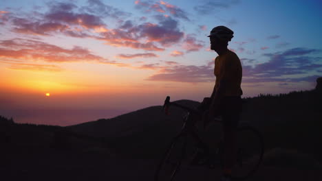 Atop-a-mountain,-athlete-in-yellow-t-shirt,-helmet,-and-gear-rests-on-bike,-taking-in-mountain-view-and-sunset-after-training