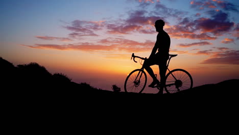 Young-sportsman,-donning-yellow-t-shirt-and-gear,-finds-solace-on-mountain-peak's-bike,-gazing-at-mountains-and-sunset-after-workout